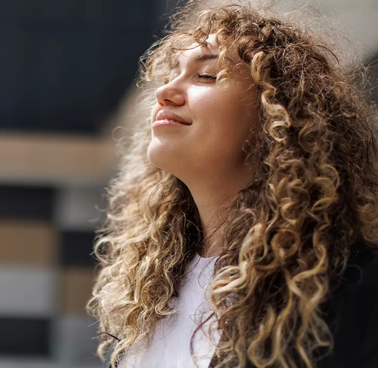 A person with curly hair smiles and closes their eyes, enjoying the sunshine outdoors.