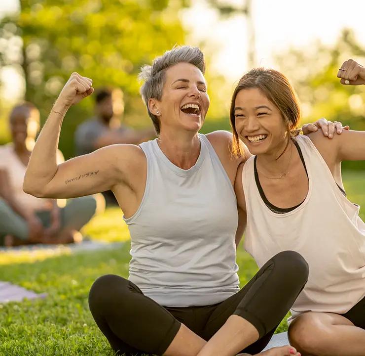 Two women sit on grass with yoga mats, smiling and flexing muscles, surrounded by people practicing yoga in a park.