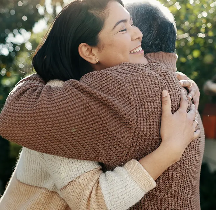 A woman and a man hug outside, smiling, with sunlight filtering through the trees in the background.