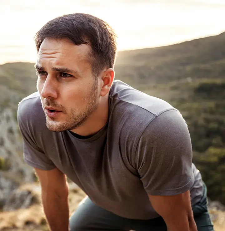Man catching breath outdoors on a rocky hill during sunset, wearing a gray shirt.