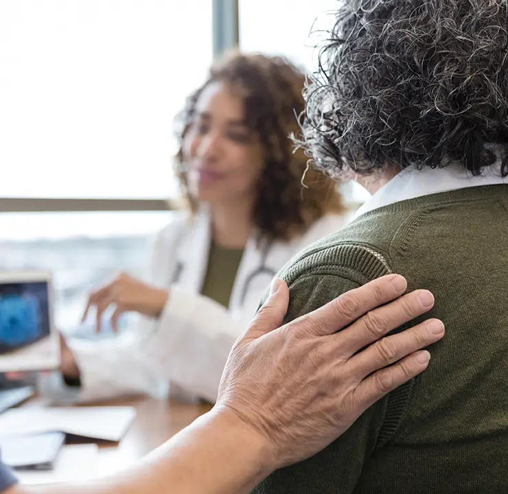 A doctor consults with two people, showing a medical image on a tablet. One person places a comforting hand on the other's shoulder.