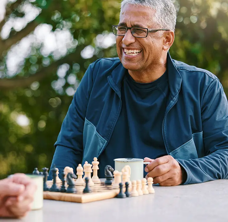 Two men smiling and talking outdoors, one wearing glasses and a dark jacket, with trees softly blurred in the background.