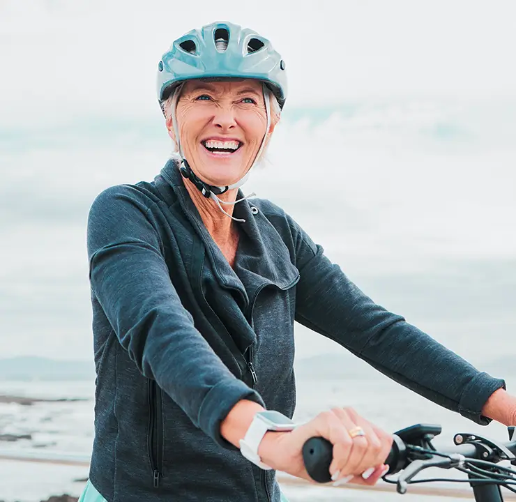 Woman smiling while wearing a helmet and dark jacket, outdoors with a cloudy sky background.