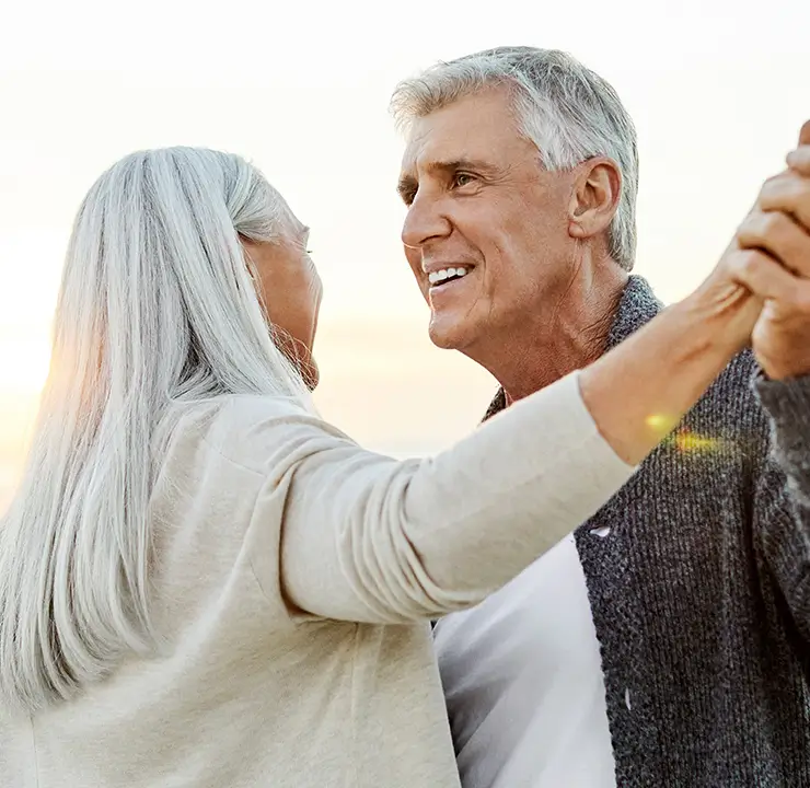An elderly couple dances joyfully at the beach during sunset.