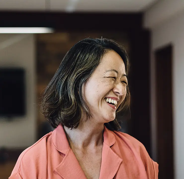 A smiling woman with shoulder-length hair wearing a pink jacket, standing indoors with a blurred TV screen in the background.