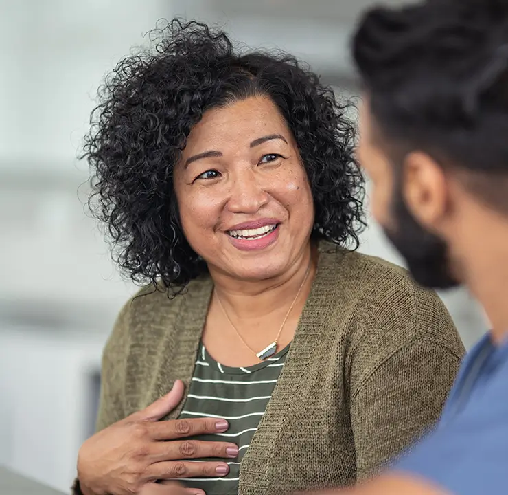 A woman in a green sweater smiles and speaks with a man in a blue shirt, who is facing her. They're sitting indoors in a cozy setting.