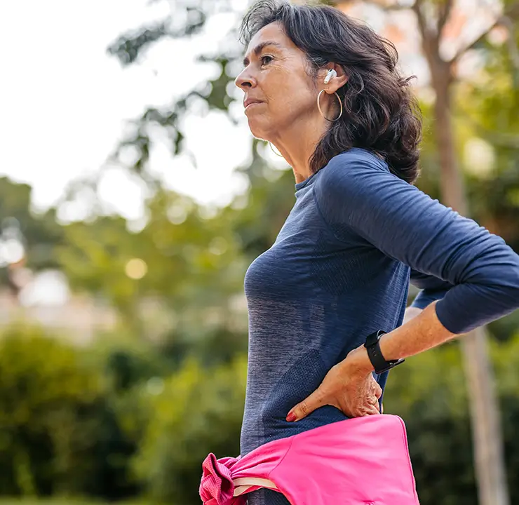 A woman with earbuds stands outdoors, looking thoughtful. She wears a long-sleeved blue shirt and has her hands on her hips. Trees are in the background.