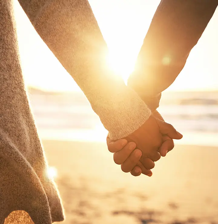 Two people holding hands at the beach during sunset, with the sun shining brightly in the background.