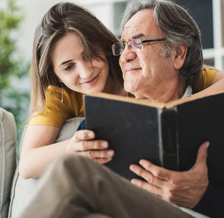 A woman leans on an elderly man's shoulder as they look at a black book together, both appearing content.
