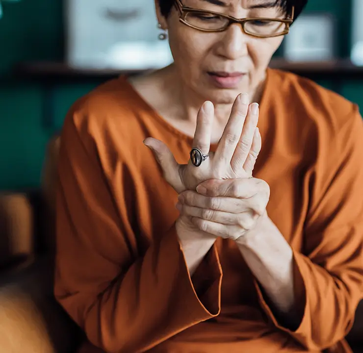 Woman in orange shirt holding her hand, appearing to experience discomfort, indoors with a blurred background.