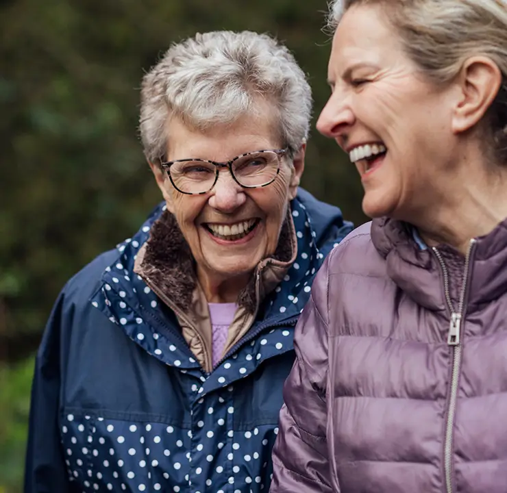Two women laughing together outdoors, both wearing jackets. The background is blurred with greenery.