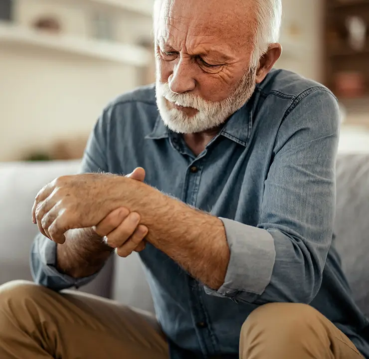 Elderly man with white beard sitting on a sofa, holding and examining his right wrist with a concerned expression.
