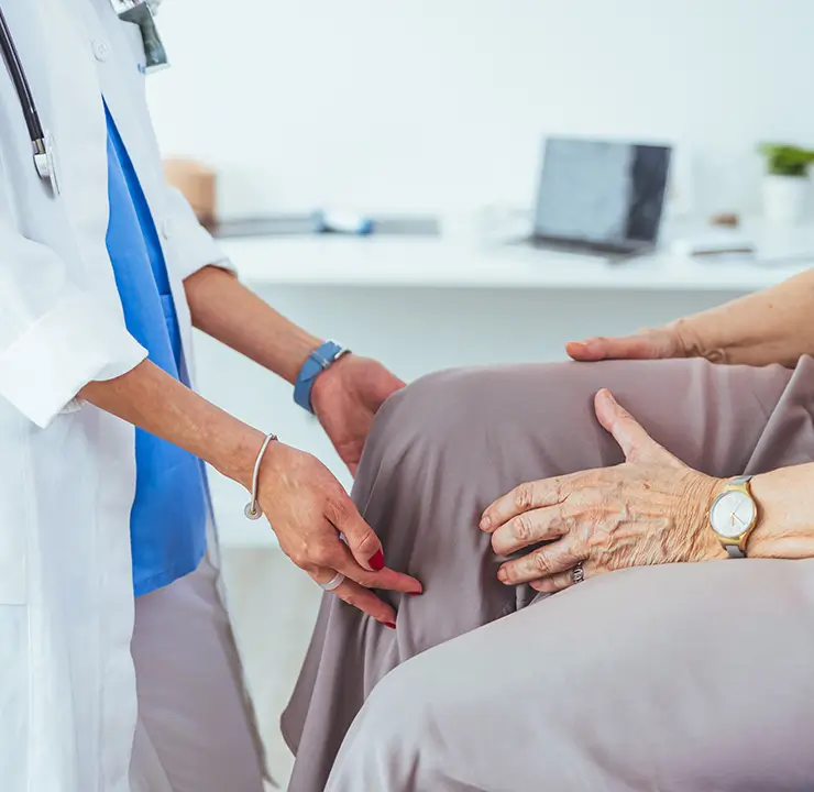 Doctor examining an elderly patient's knee, pointing and discussing the condition, with a medical office in the background.