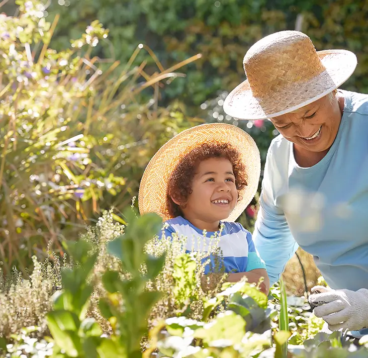 A smiling woman and child wearing straw hats enjoy gardening together in a sunny garden.