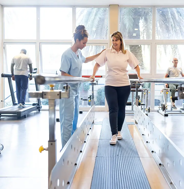 A physical therapist assists a woman walking on a parallel bar in a rehabilitation center with others exercising in the background.