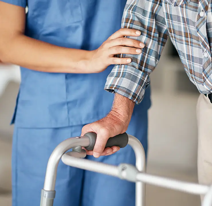 A caregiver supports a person using a walker. The caregiver, in blue scrubs, holds the person's arm for assistance indoors.