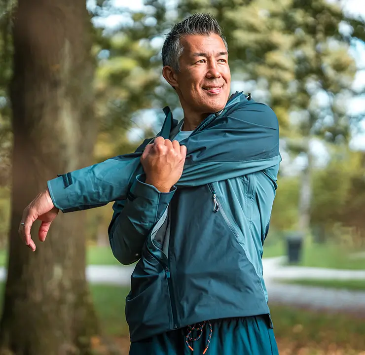 Man in a blue jacket stretches his arm outdoors, surrounded by trees and greenery.