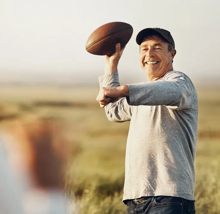An older man in a cap smiles while throwing a football outdoors, with a blurred figure in the foreground.