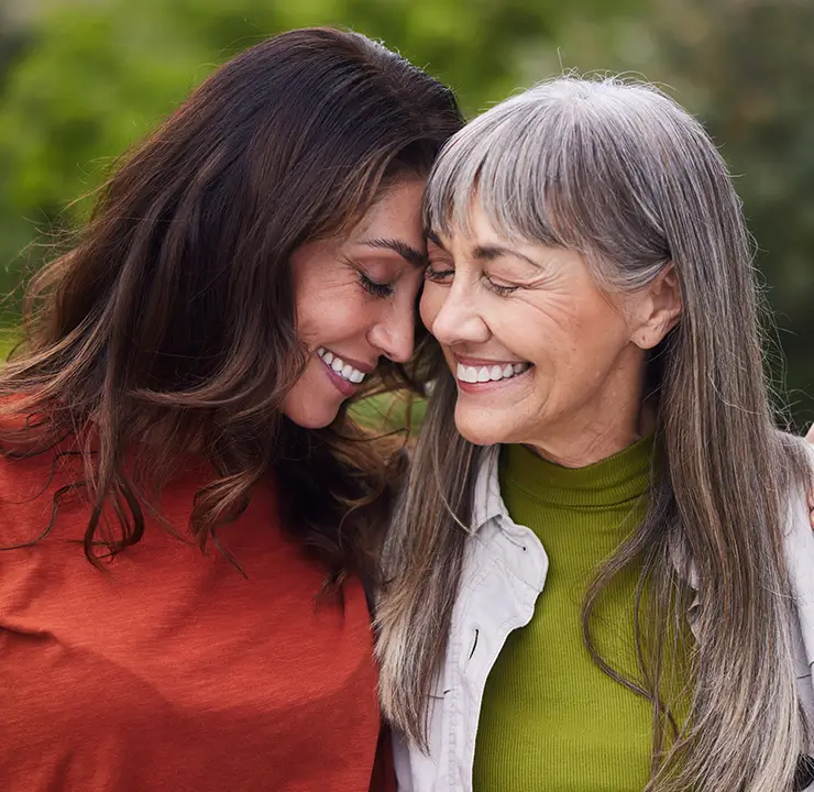 A younger woman in an orange shirt and an older woman in a green sweater share a joyful embrace, smiling with their heads touching.