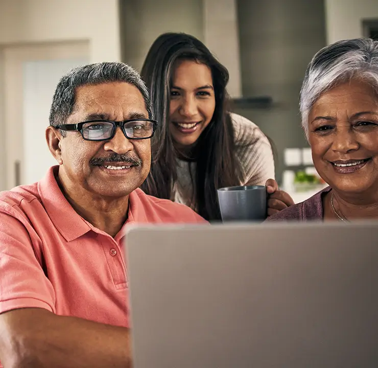 Three people smiling and looking at a laptop screen. The older man and woman are seated, and the younger woman stands behind them holding a mug.