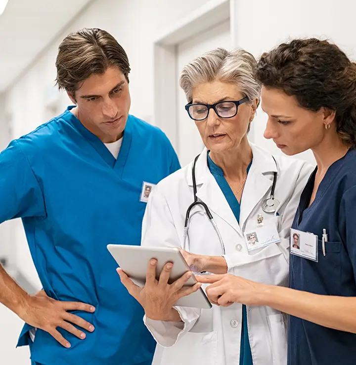 Three medical professionals in scrubs and lab coats discuss information on a tablet in a hospital corridor.
