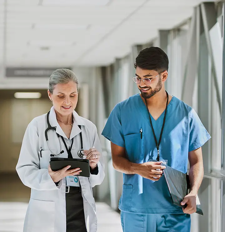 Two healthcare professionals walking in a hallway; a female doctor with a tablet and a male nurse in blue scrubs holding a folder, both smiling.