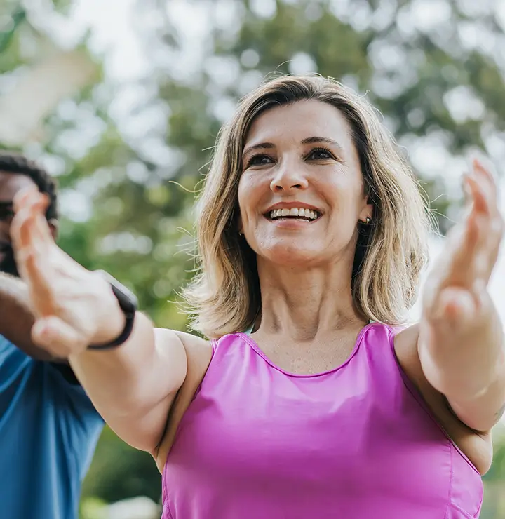 Two adults smiling and stretching their arms forward outdoors, wearing casual athletic wear. Trees are blurred in the background.
