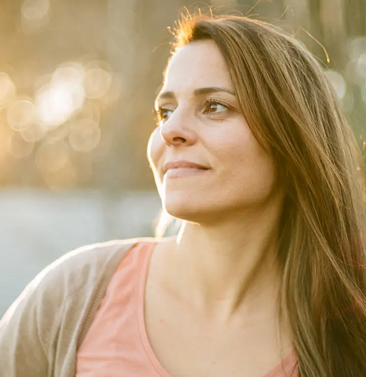 Woman with long hair looks to the side, smiling softly, with sunlight creating a warm, blurred background.