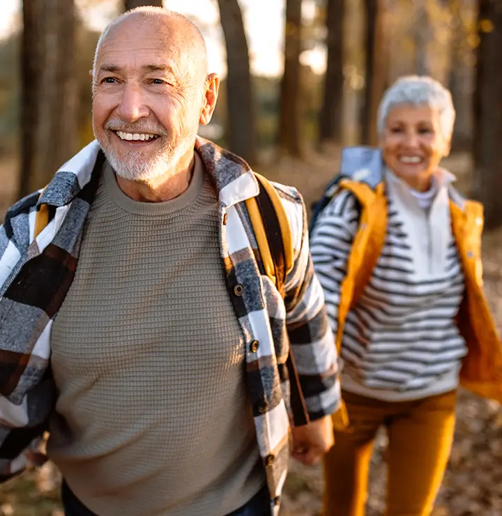 An elderly couple smiles while hiking through a sunlit forest in autumn, wearing casual outdoor clothing with backpacks.