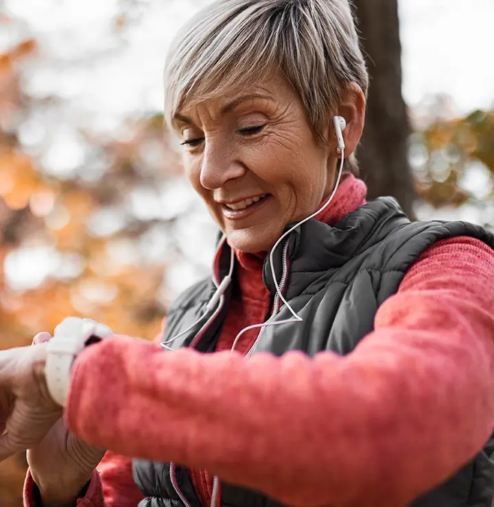 A woman wearing earbuds checks her smartwatch outdoors, surrounded by autumn leaves.