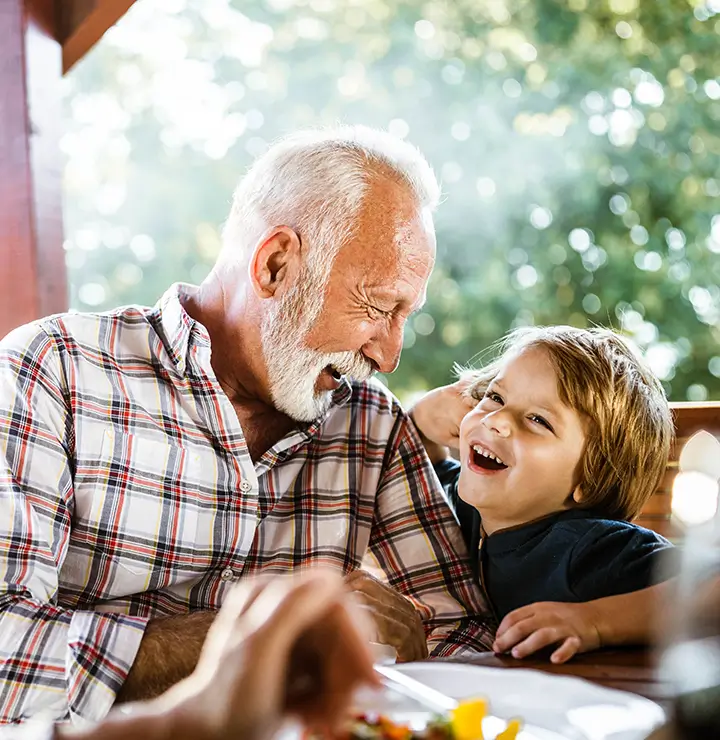 A joyful elderly man and a laughing child share a cheerful moment at a sunlit wooden table outdoors.