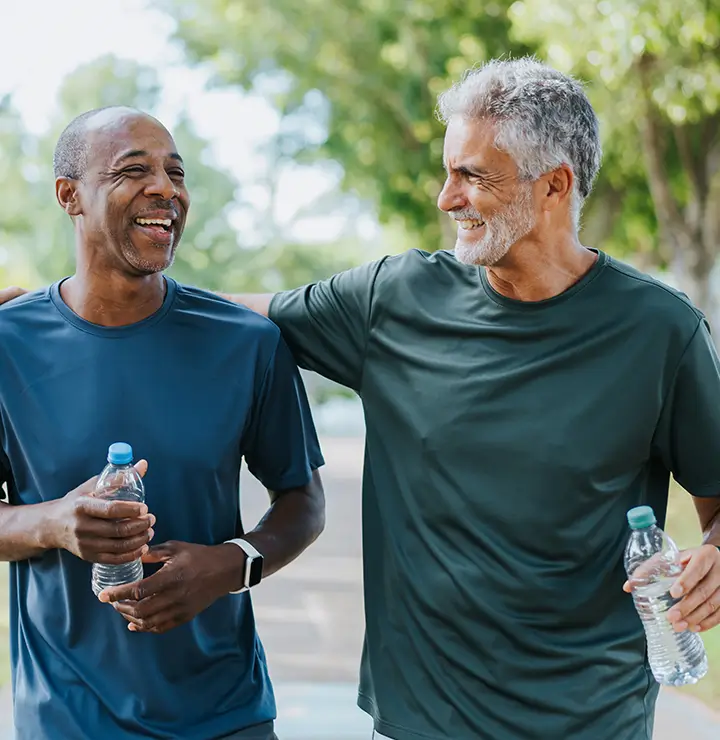 Two men in athletic shirts smiling and walking outdoors, one holding a water bottle. Trees and greenery are visible in the background.