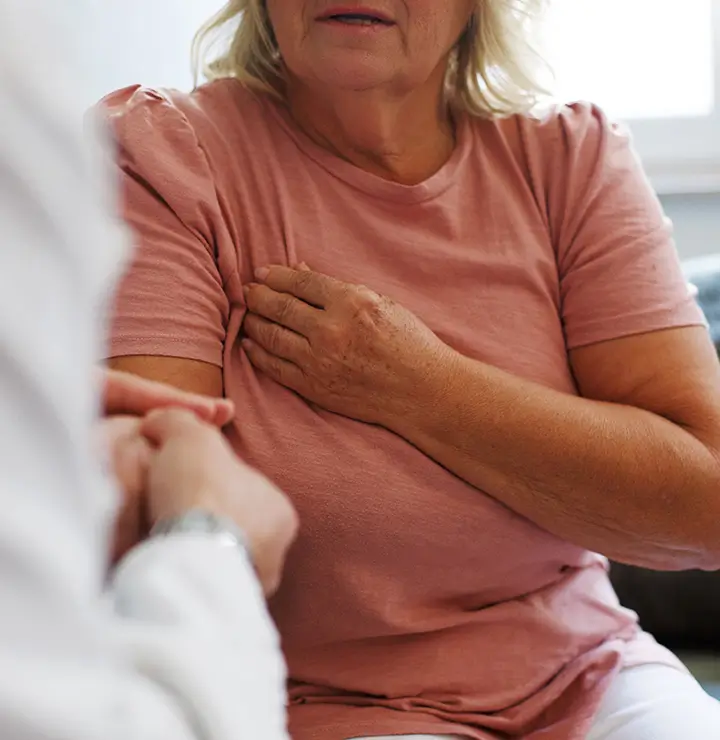 A woman in a pink shirt holds her chest, appearing distressed, while speaking to a doctor.