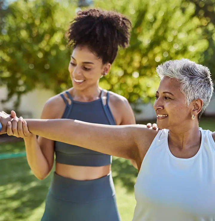 A woman helps another woman exercise with a dumbbell in a sunny outdoor setting.