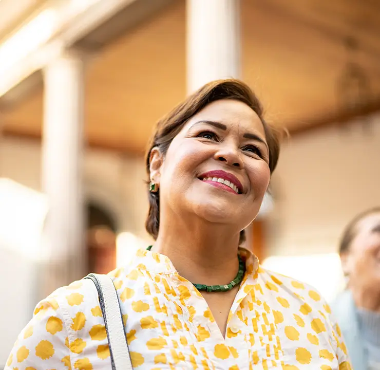Two women smiling outdoors, one in focus wearing a yellow patterned top. Bright, sunny day with buildings in the background.