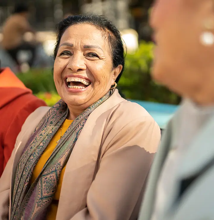 Three women sitting on a bench, smiling and laughing outdoors on a sunny day.