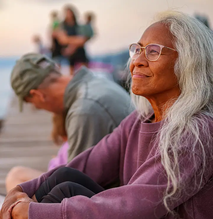 Elderly woman with long gray hair and glasses smiling peacefully by a lake; blurred background with people and trees during sunset.