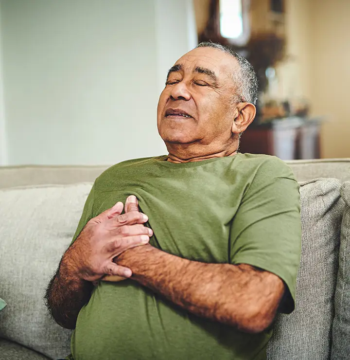 Elderly man sitting on a sofa, clutching his chest with a pained expression, wearing a green shirt.
