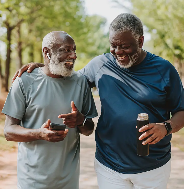 Two older men with gray beards are smiling and walking outside. One holds a smartphone, and the other carries a thermos. Trees in the background.