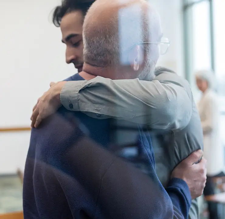 Two people embracing warmly in a well-lit room, with blurred background elements suggesting a comforting or supportive environment.