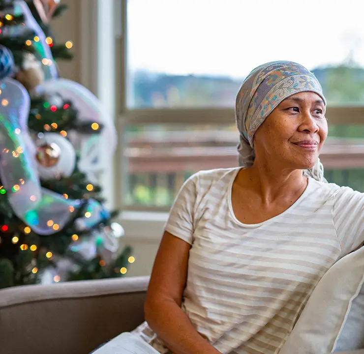 A woman in a headscarf sits on a couch near a decorated Christmas tree, looking out the window with a gentle smile.