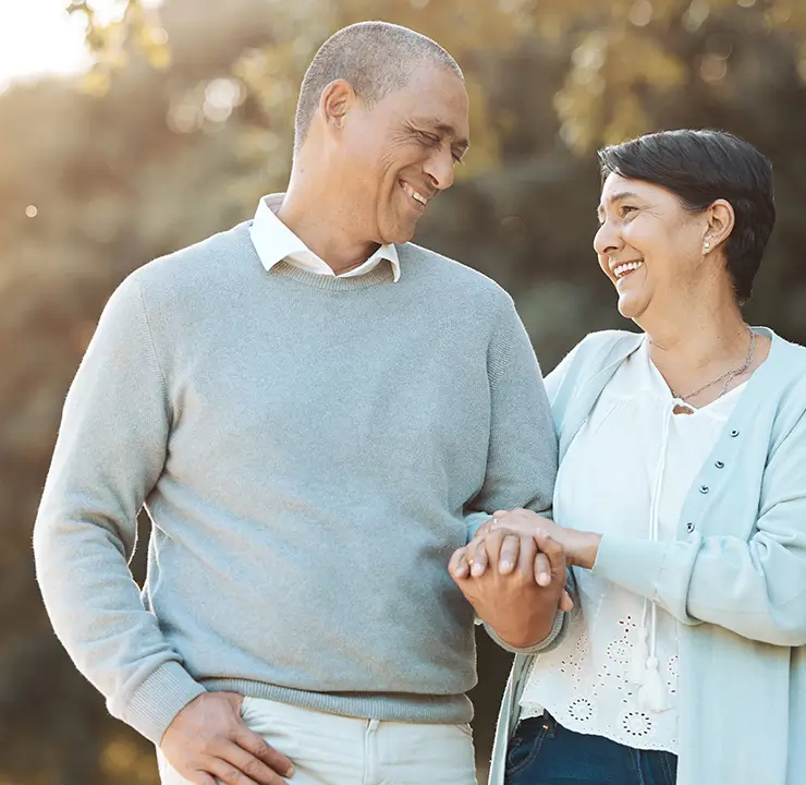 A smiling couple walks outdoors, looking at each other lovingly, with sunlight filtering through trees in the background.