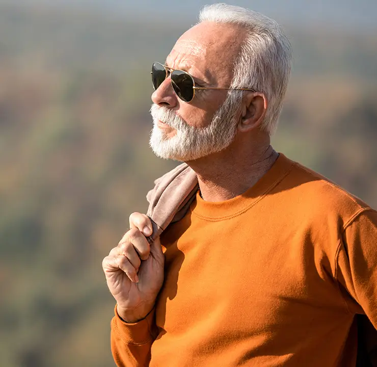 Elderly man with sunglasses and a white beard gazing at the sky, wearing an orange shirt, with a jacket over his shoulder, in an outdoor setting.