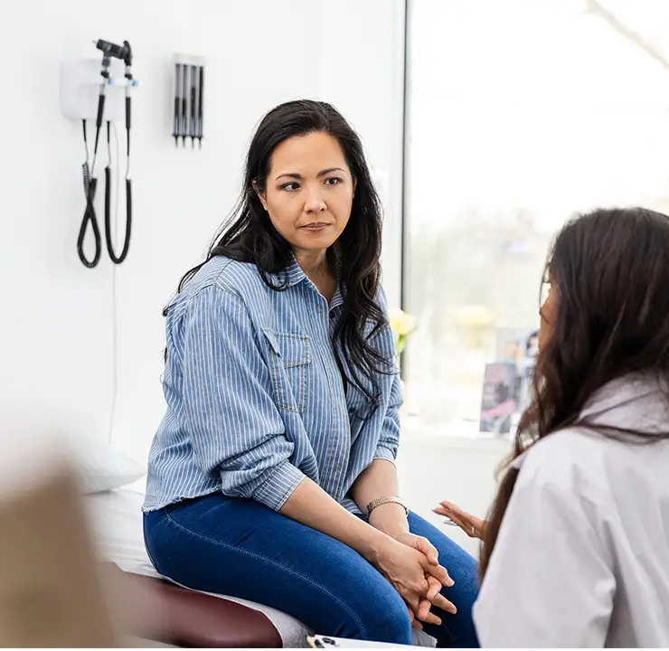 A woman in a striped shirt is sitting on an exam table, attentively listening to another woman in a medical office.