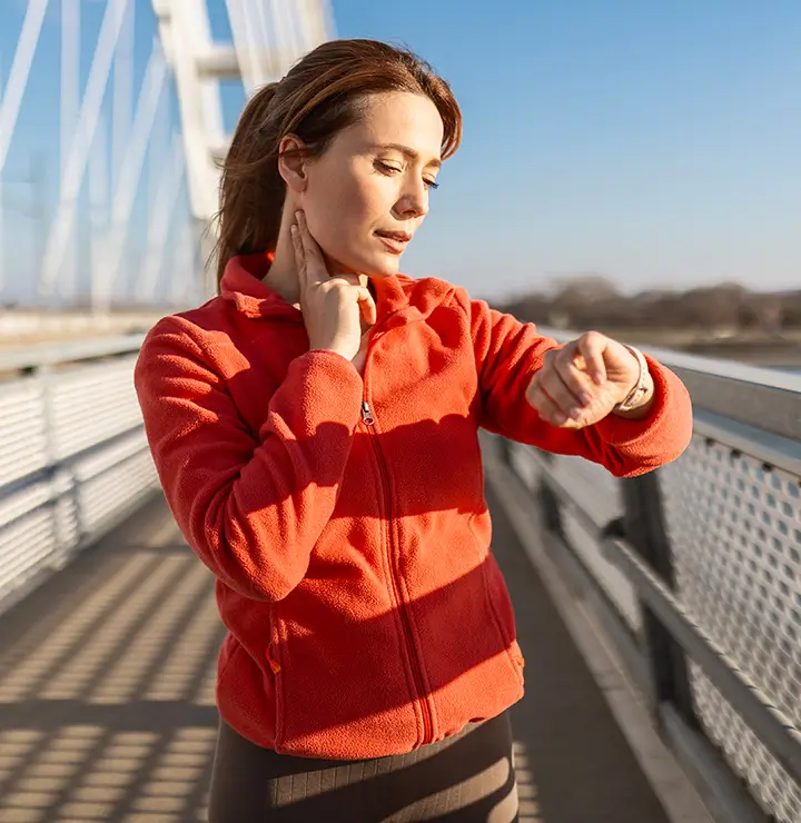 Woman in a red jacket checks her pulse and looks at her smartwatch while standing on a bridge under a clear blue sky.