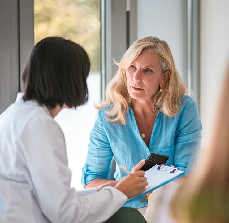 Two women having a serious conversation by a window; one with shoulder-length black hair, the other with long blonde hair wearing a blue shirt.