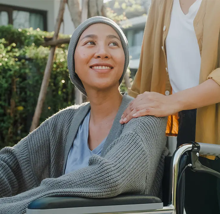 A person in a wheelchair smiles while another stands beside them, placing a reassuring hand on their shoulder, outdoors in a garden setting.