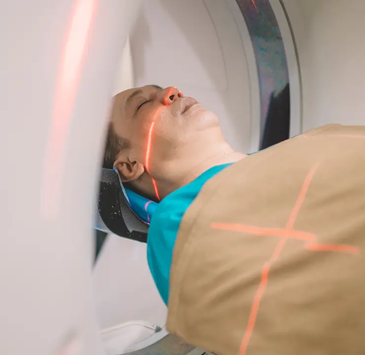 Person undergoing a CT scan, lying on a table with head inside the scanner, red alignment lights visible on the face.