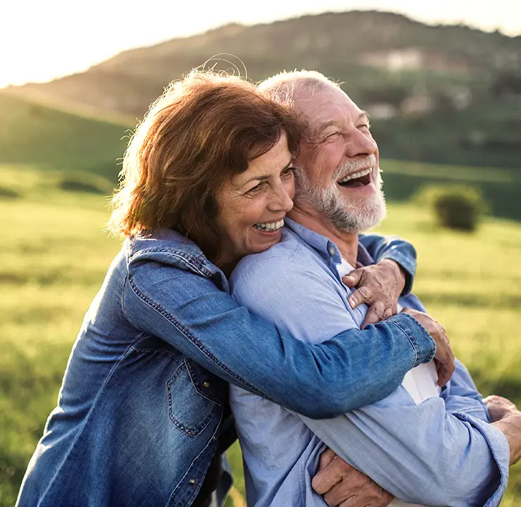 An elderly couple laughing and hugging in a sunlit green field, with hills in the background.