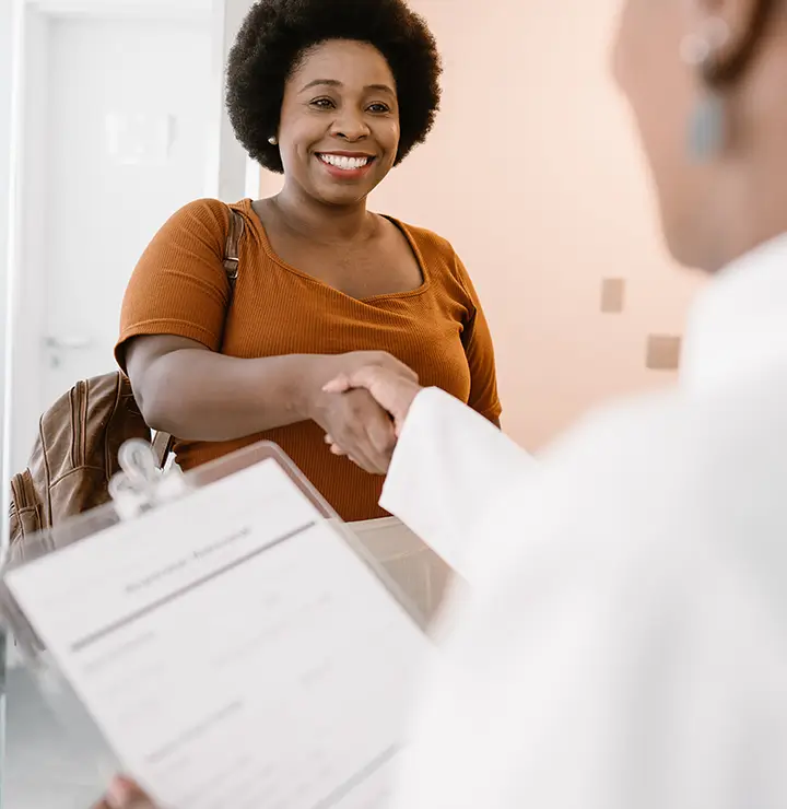 Woman in orange shirt smiling and shaking hands with a person in white attire in a bright room.